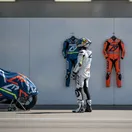 A MotoGP rider in a pit garage contemplates a choice between a blue Yamaha racing suit and an orange Honda one.