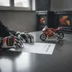 A close-up view of a rider's gloved hands signing an official HRC contract, with a model Honda race bike blurred in the background.