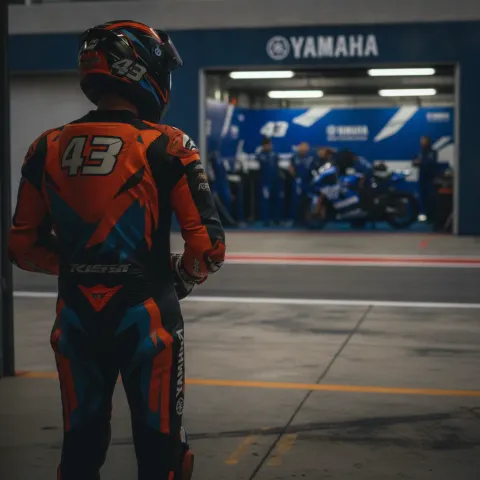 A MotoGP rider in orange leathers contemplates a move to a rival manufacturer's blue-liveried team.