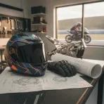 A racing helmet and gloves lie on a table next to technical drawings of a motorcycle engine, symbolising a rider's future move.