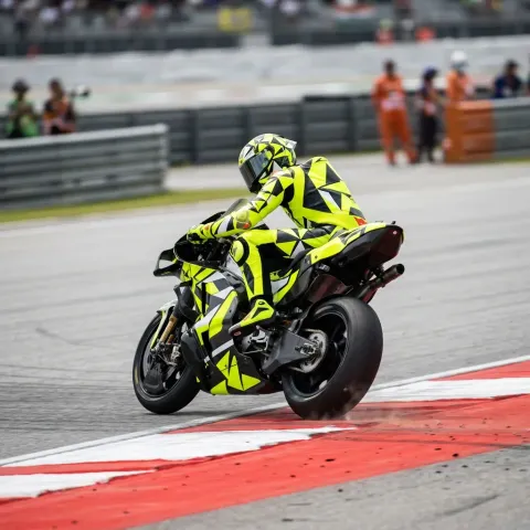 A MotoGP rider in an invented yellow-and-black livery accelerates out of a corner over a red-and-white kerb.