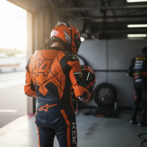 A replacement MotoGP rider stands in a pit garage, holding his helmet and preparing to ride.