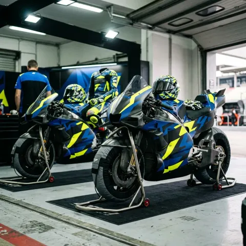 Two identical MotoGP race bikes being prepared by mechanics in a team's pit garage, illustrating the team effort.